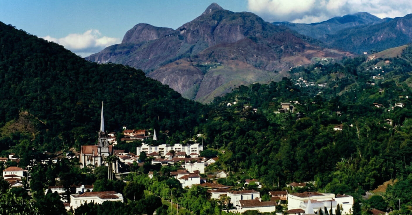 Vista panorâmica da cidade de Petrópolis, RJ, situada em um vale cercado por montanhas cobertas pela Mata Atlântica. Em destaque no centro, a Catedral de São Pedro de Alcântara com sua torre neogótica, rodeada por casarões históricos e vegetação densa sob céu azul.